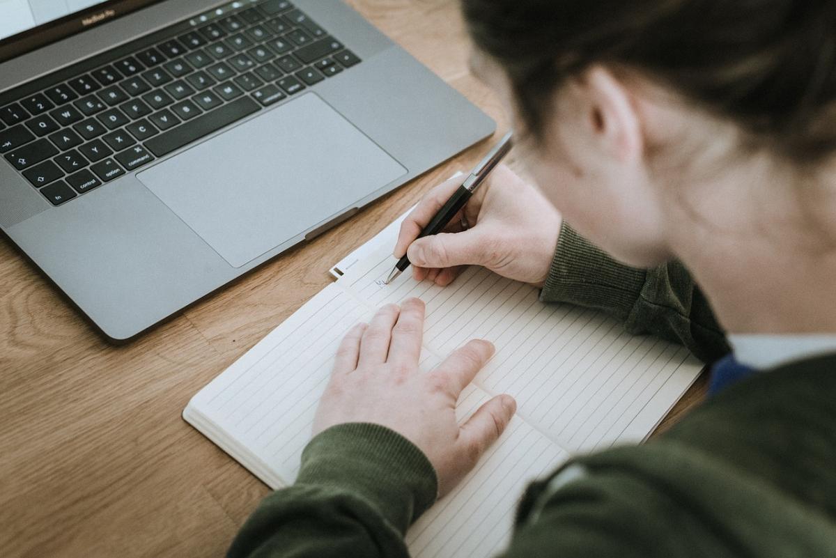 Laptop on table with person writing in a notebook